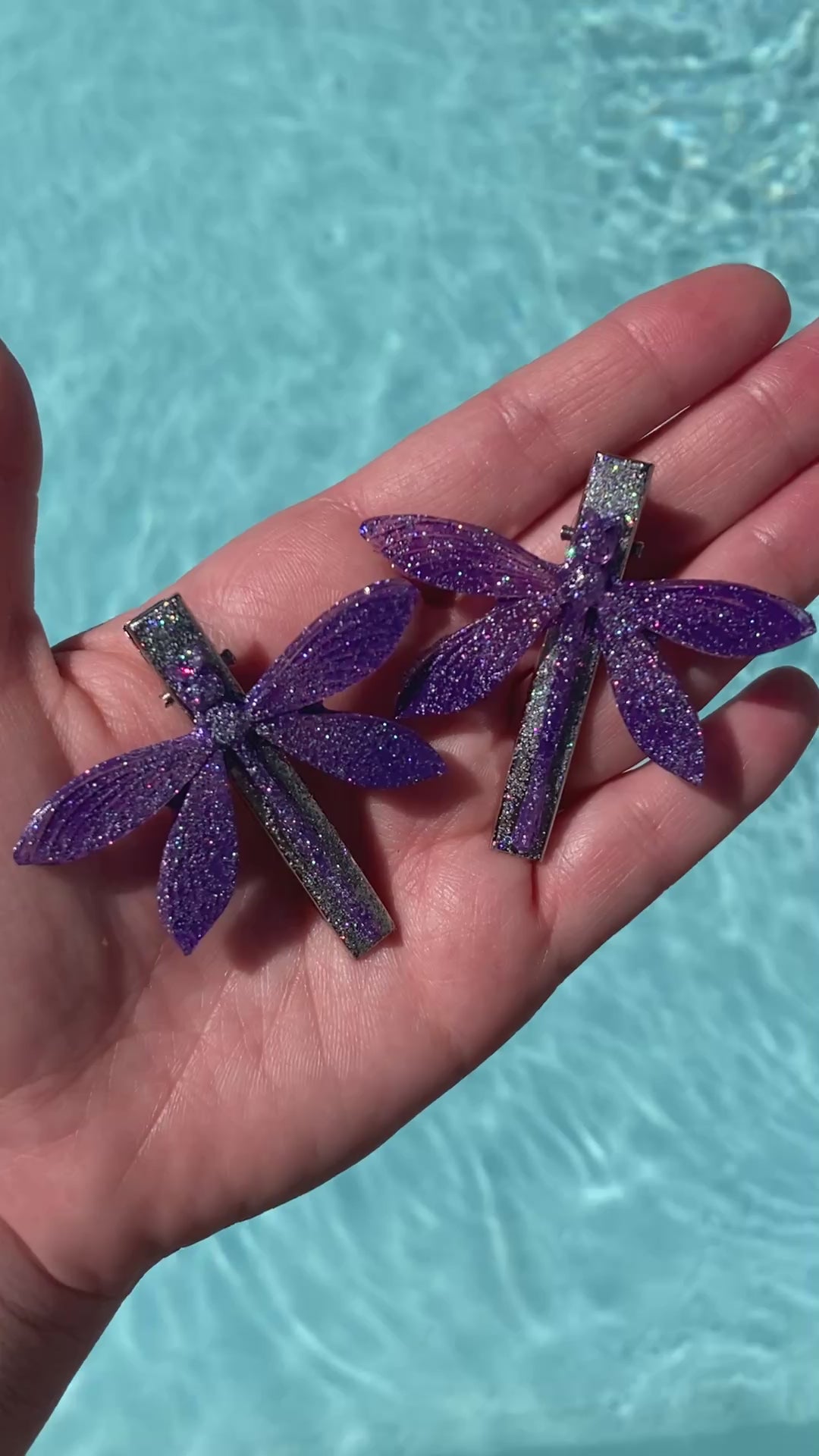 Video shows a set of two purple dragonfly hair clips in front of a pool