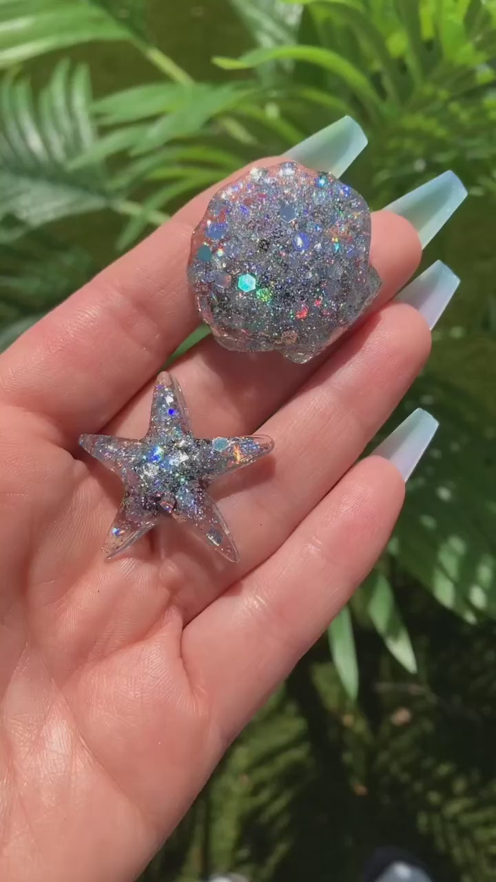 Video shows two shell shaped sequin car vent clips in a hand in front of a plant 