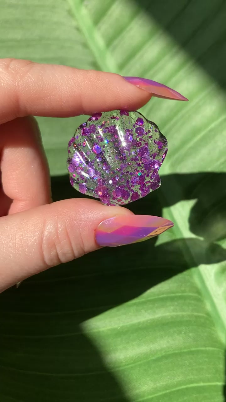 Video shows a resin shell with purple sequins being held by a manicured hand in front of a leafy background 