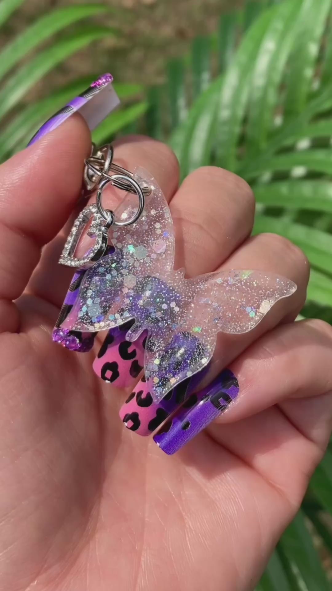Video shows a sequin butterfly keychain with a rhinestone heart charm in a manicured hand in front of a artificial plant.