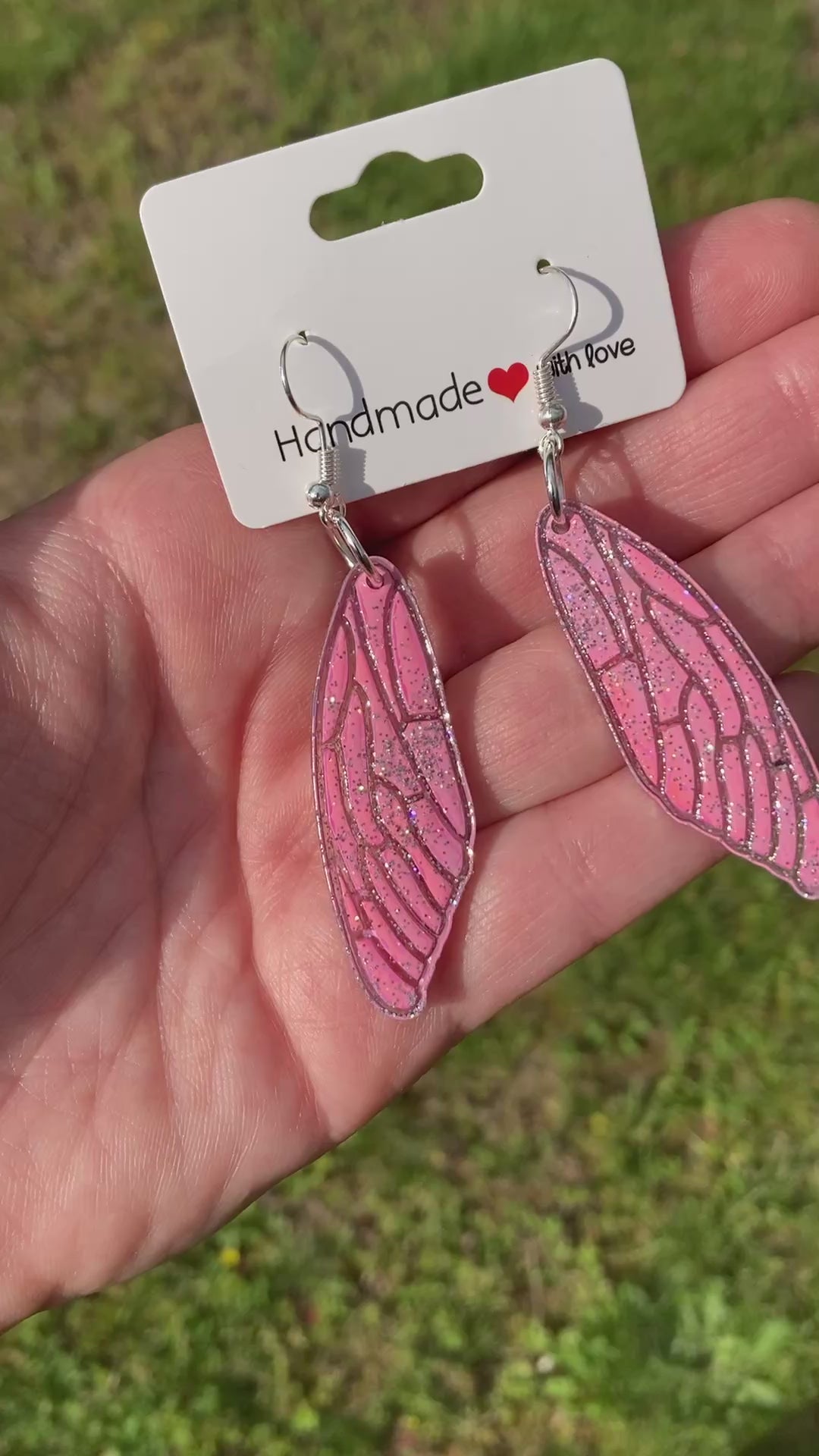 Video shows a pair of pink butterfly wing earrings in front of grass being held by a hand 