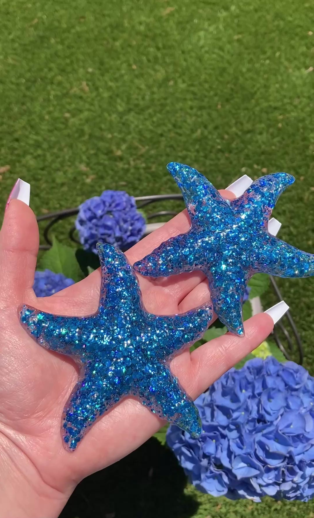 Video shows two resin blue sequin starfish in front of blue flowers 