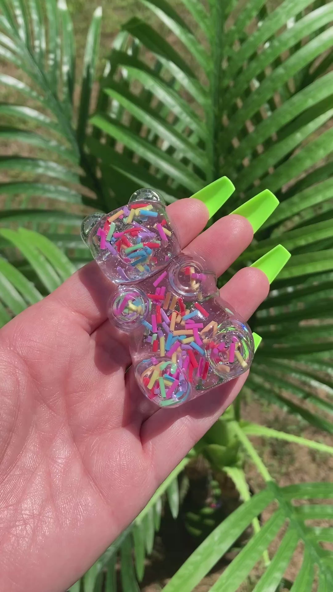 Video shows a manicured hand holding a resin teddy bear figurine with colorful sprinkles in front of a artificial plant.