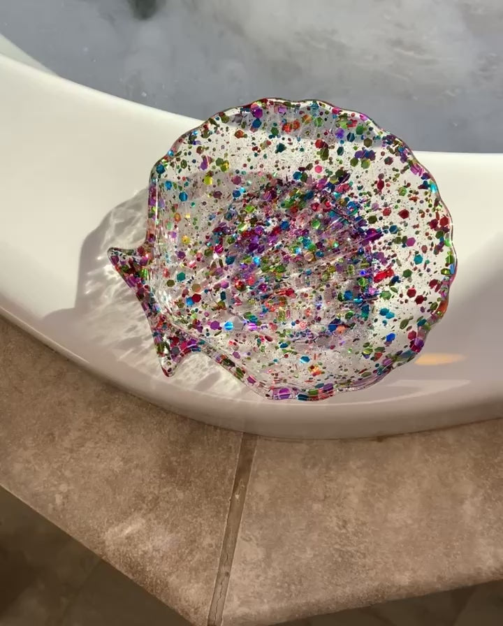 Video shows a rainbow colored sequin seashell soap dish on the edge of a tub