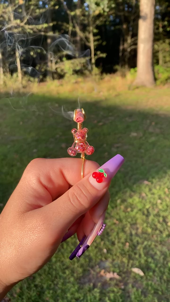 The video shows a pink sequin teddy bear smoke ring lit with a faux joint in a nature setting. 
