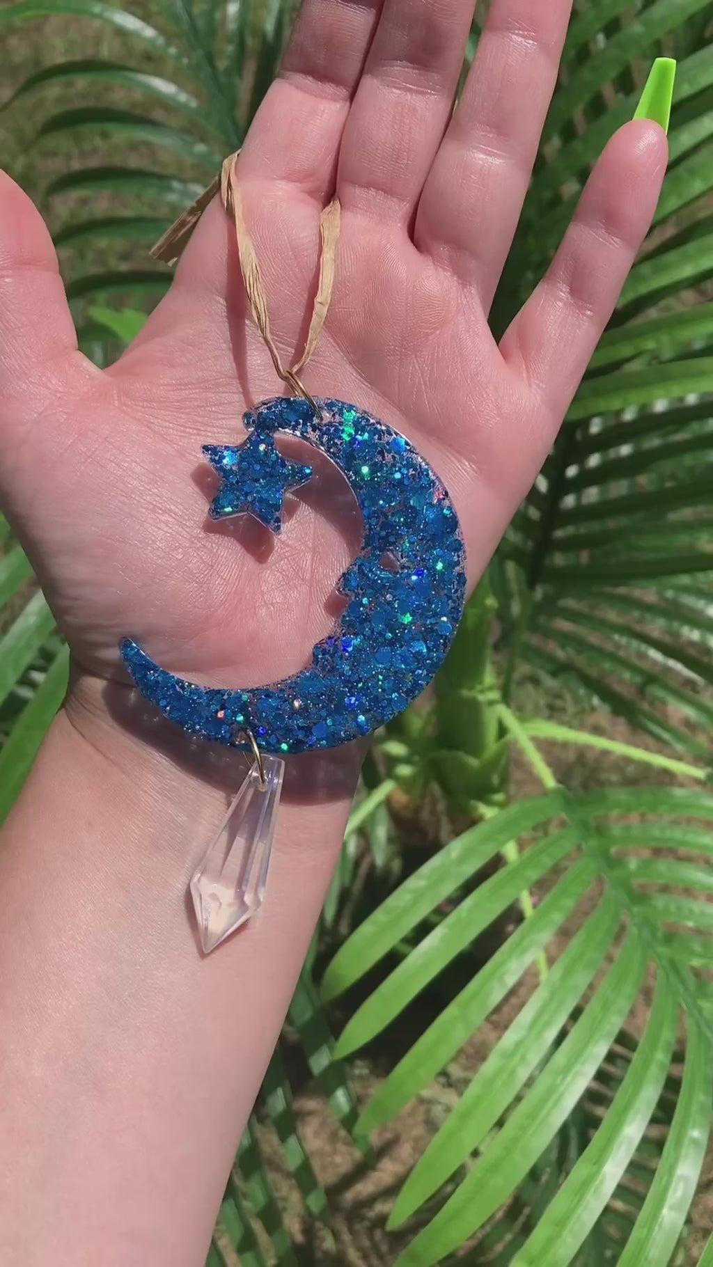 Video shows a manicured hand holding a blue sequin crescent moon with a crystal dangling sun catcher in front of a artificial plant.