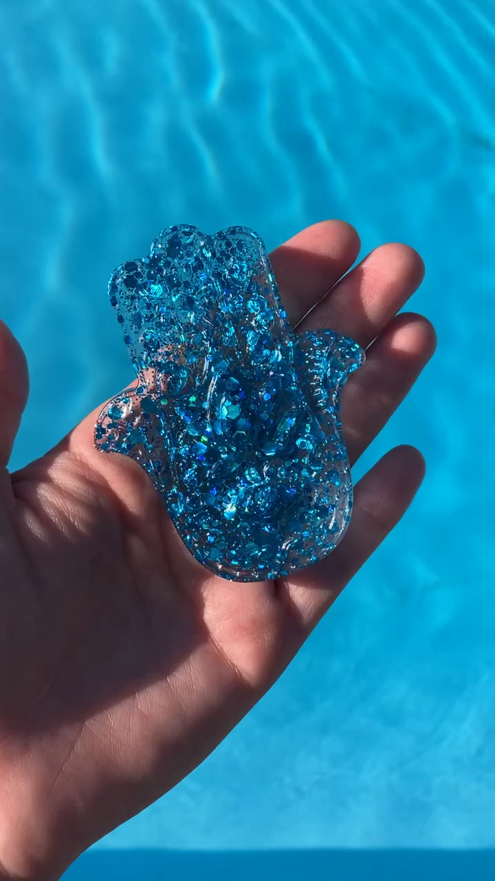 Video shows a hand holding a blue sequin hamsa resin hand decor above a pool