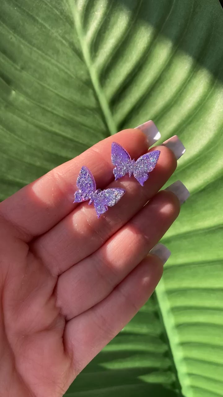 The video shows a hand holding a pair of butterfly stud purple earrings 