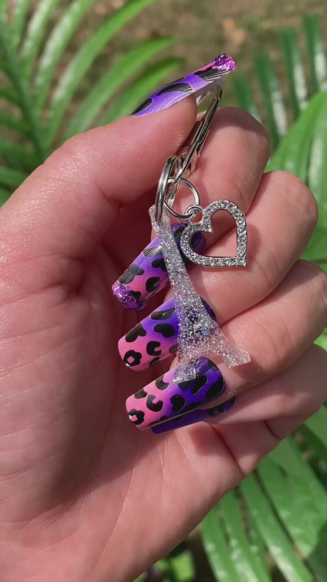Video shows a manicured hand holding an Eiffel Tower keychain with a rhinestone heart charm in front of an artificial plant.