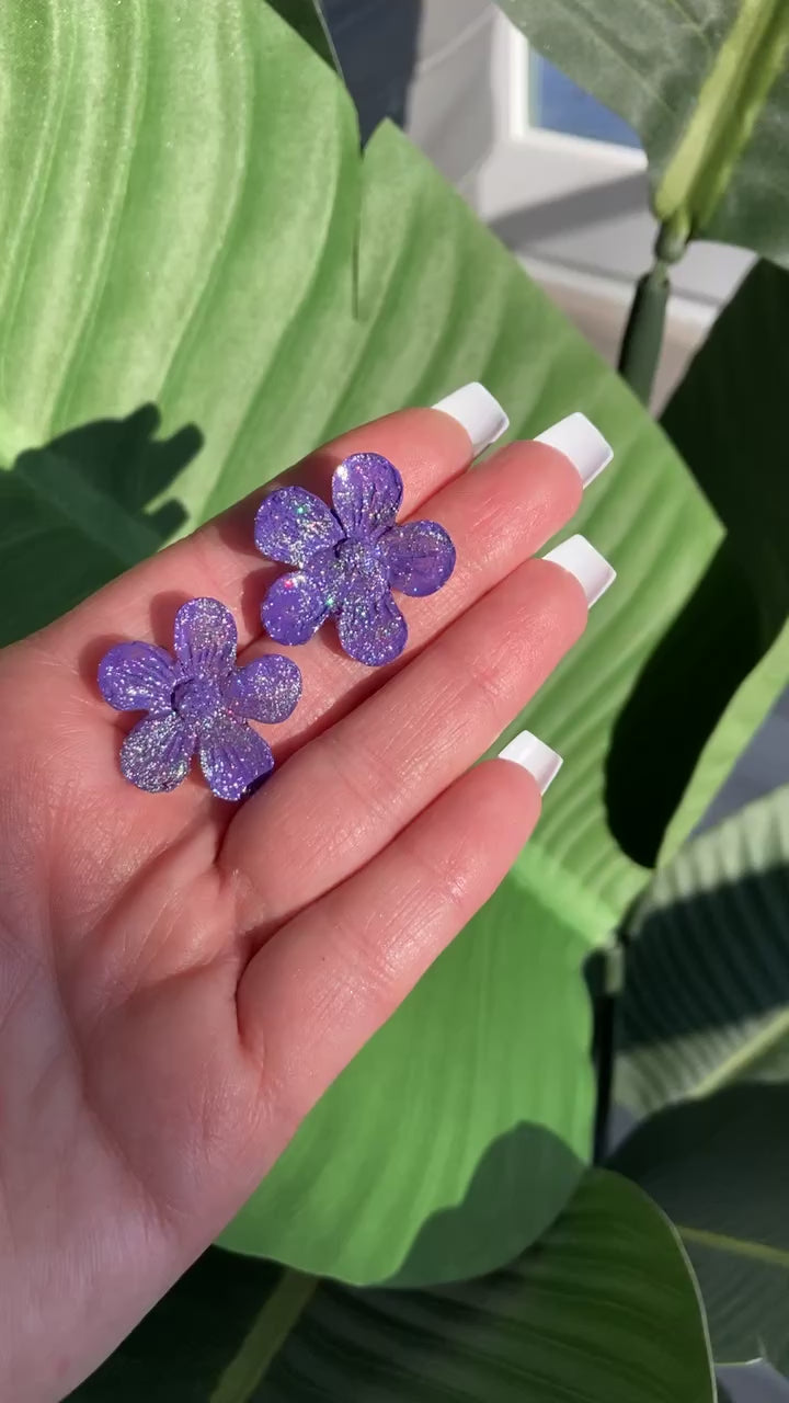 Video shows a hand holding a pair of purple sparkle flower earrings 