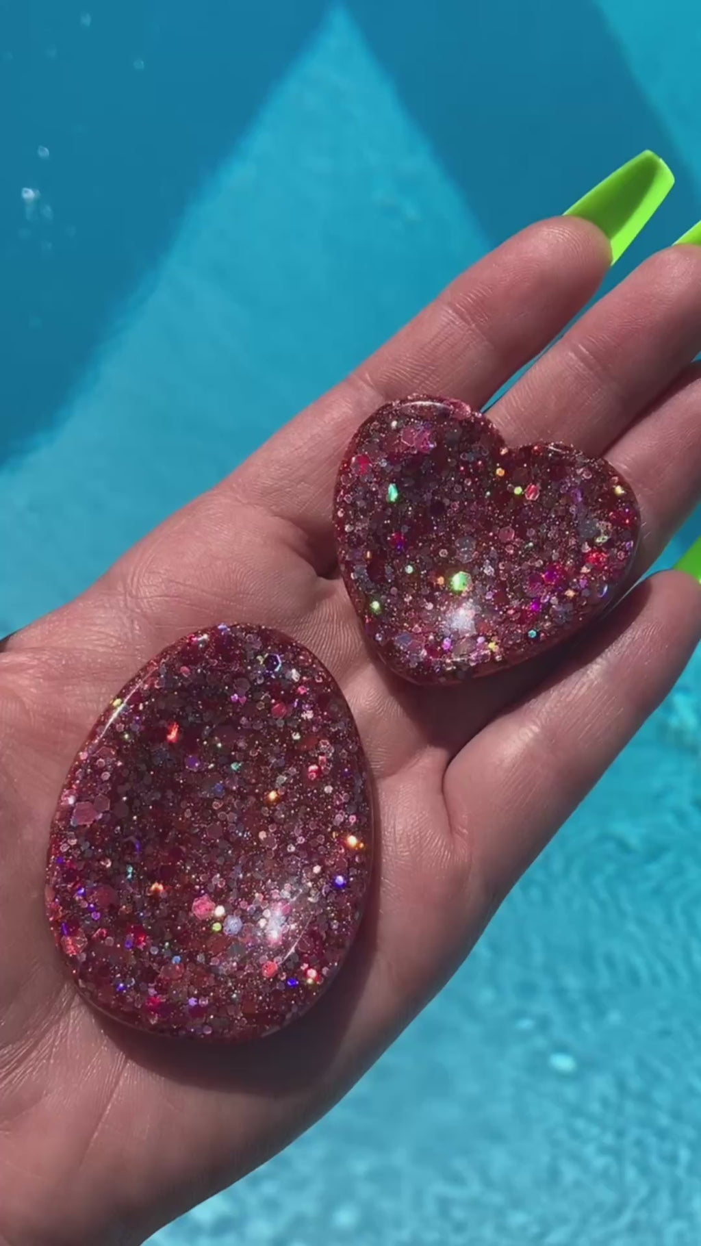 Video shows a manicured hand holding two pink sequin anxiety tokens, one heart shaped and the other oval shaped, in front of pool water.