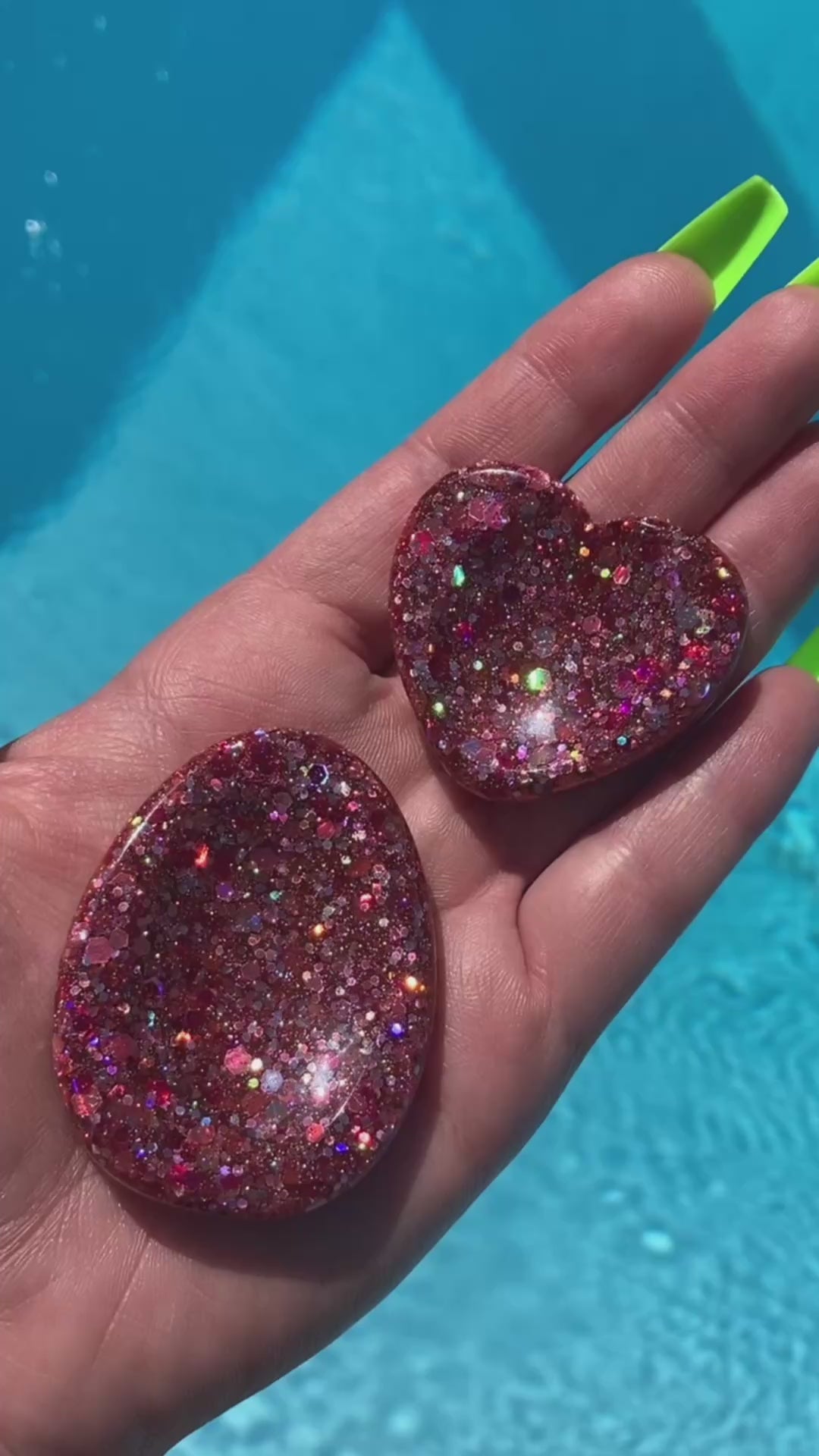 Video shows a manicured hand holding two pink sequin anxiety tokens, one heart shaped and the other oval shaped, in front of pool water.