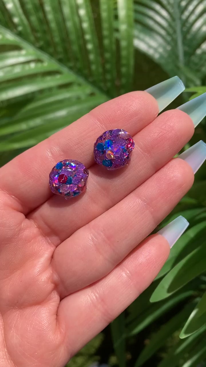 Video shows a hand holding a pair of mermaid colored shell stud earrings in front of a artificial plant.