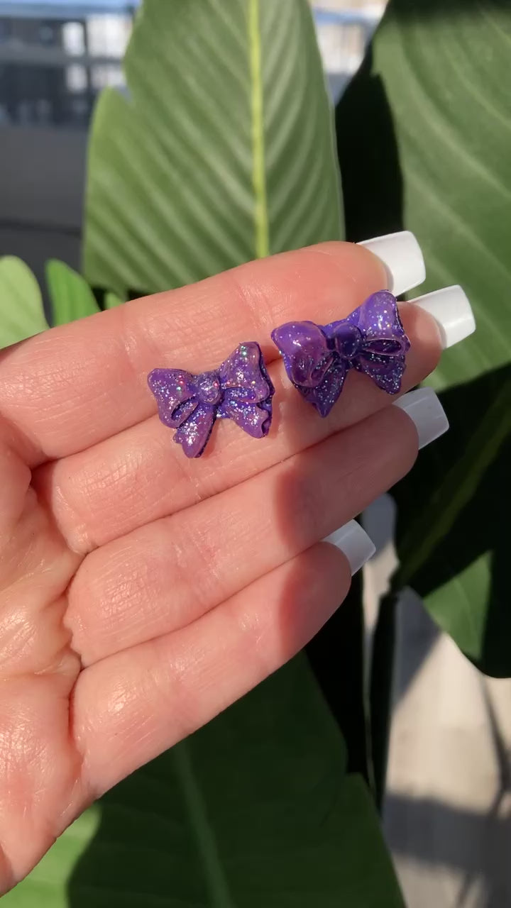 The video shows a hand holding a pair of purple glitter bow stud earrings 