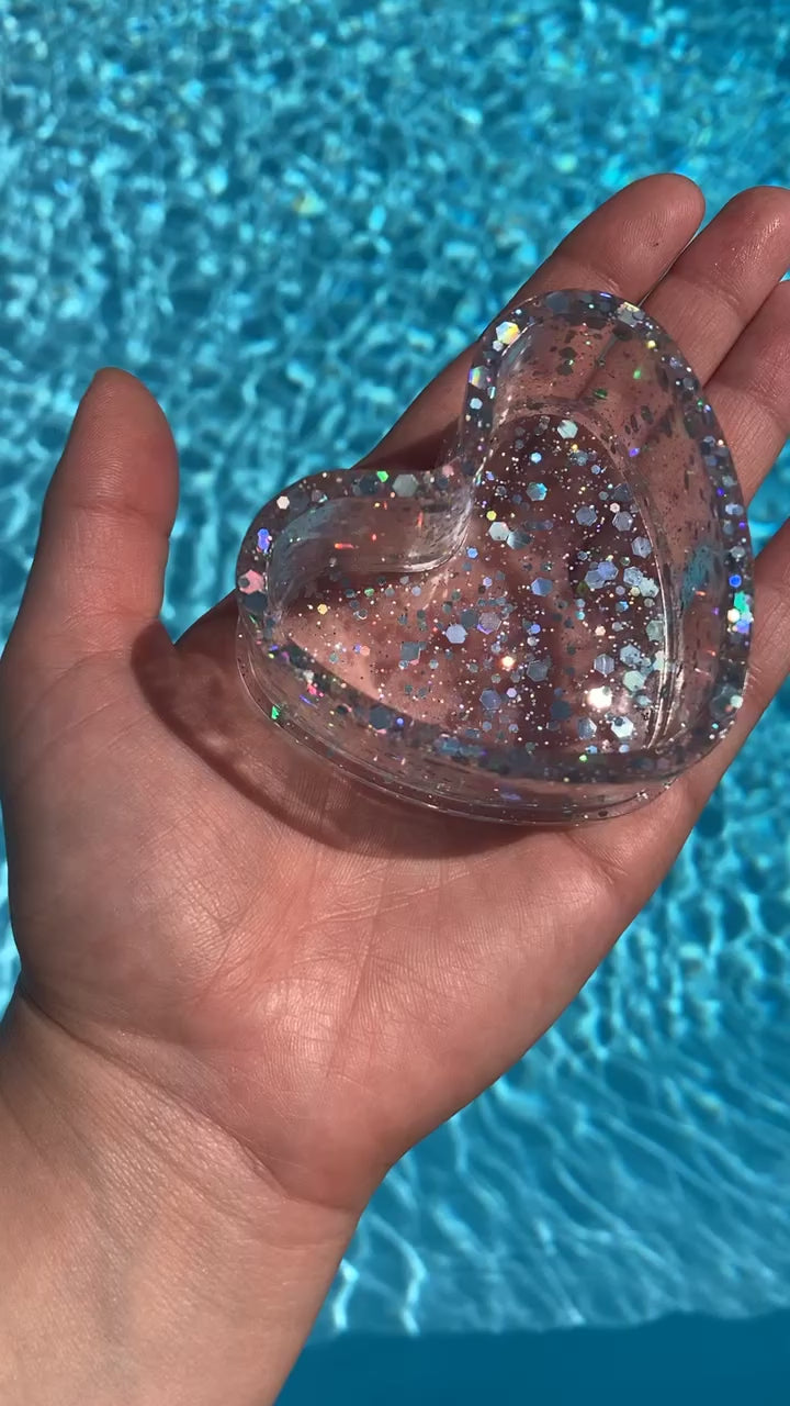 The video shows a hand holding a sequin heart dish in front of water.