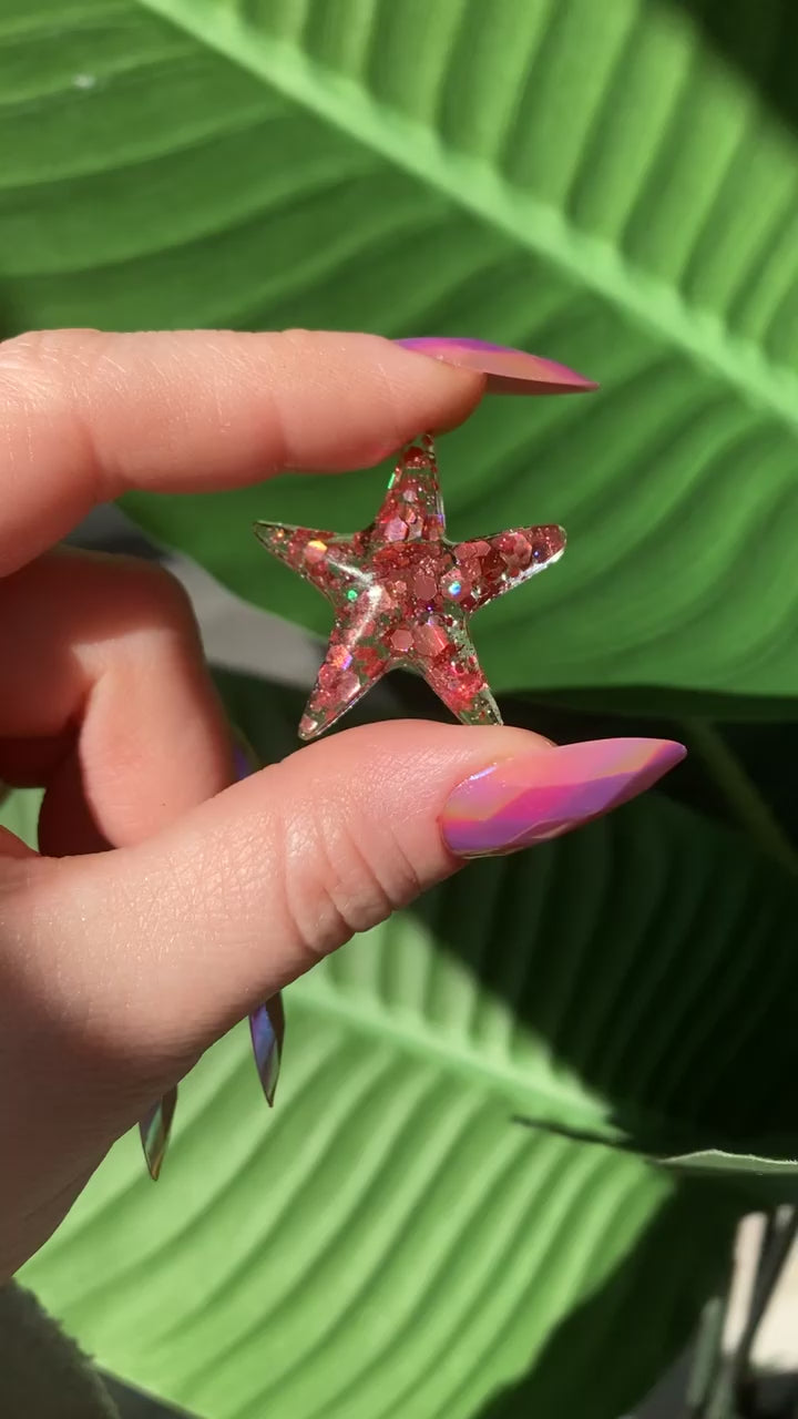 Video shows a pink resin starfish being held by a manicured hand in front of a leafy background 