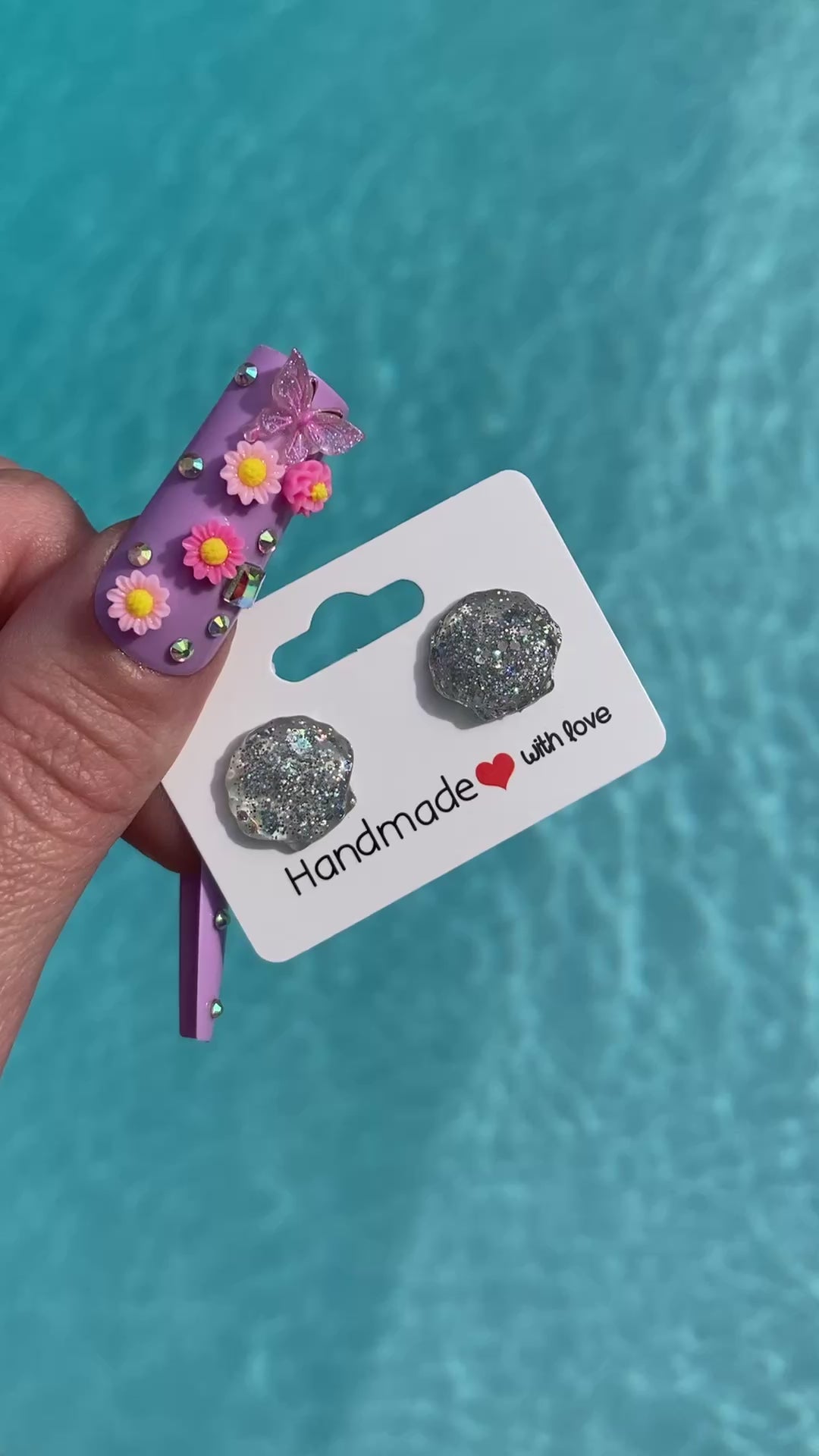 Video shows a pair of silver sequin shell earrings on a card that says “handmade with love” in front of a pool.