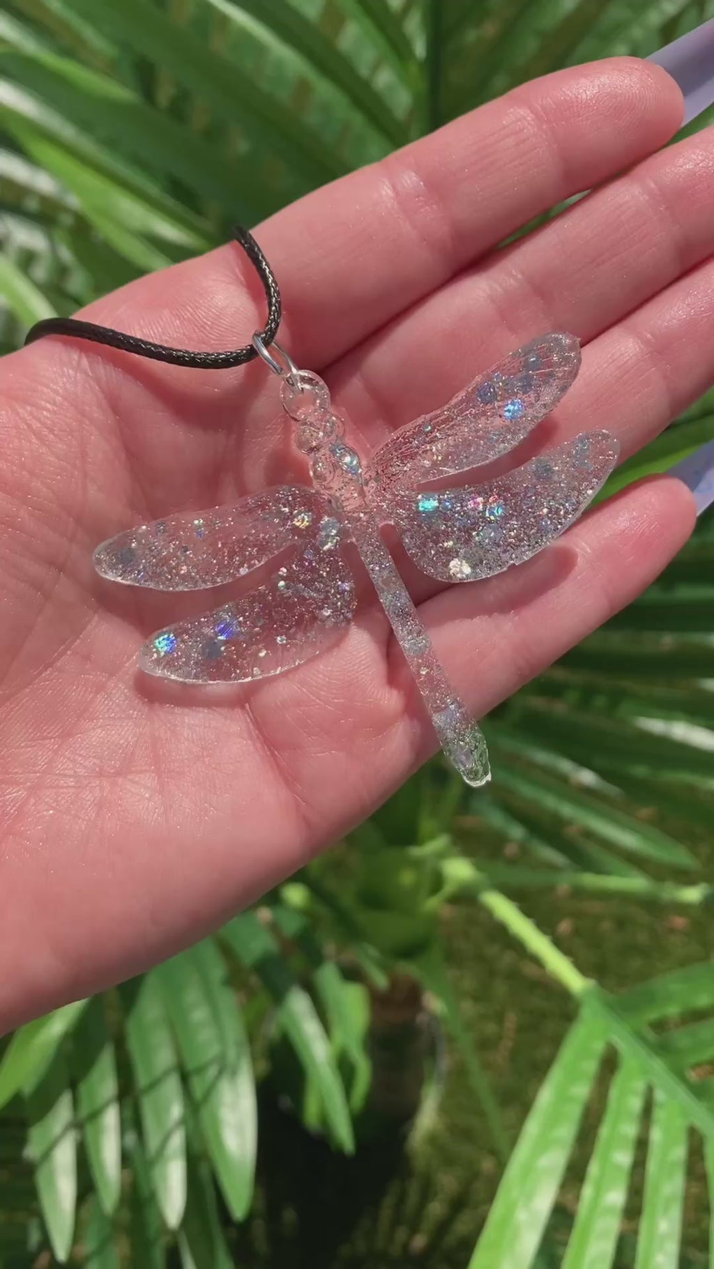 Video shows a glittery resin dragonfly necklace in front of a plant
