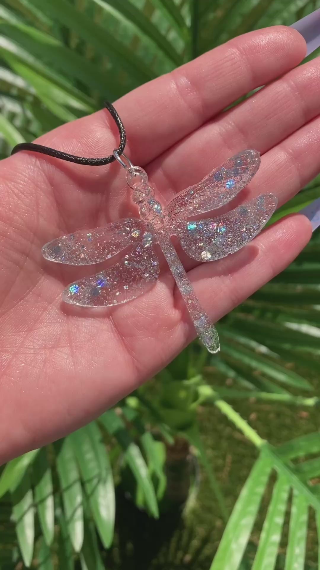 Video shows a glittery resin dragonfly necklace in front of a plant