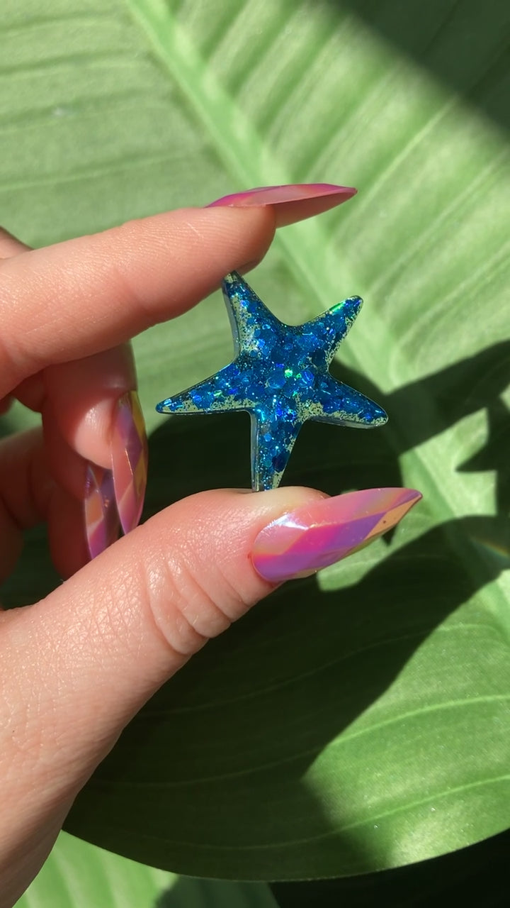 Video shows a resin blue starfish in a manicured hand in front of a leafy background 