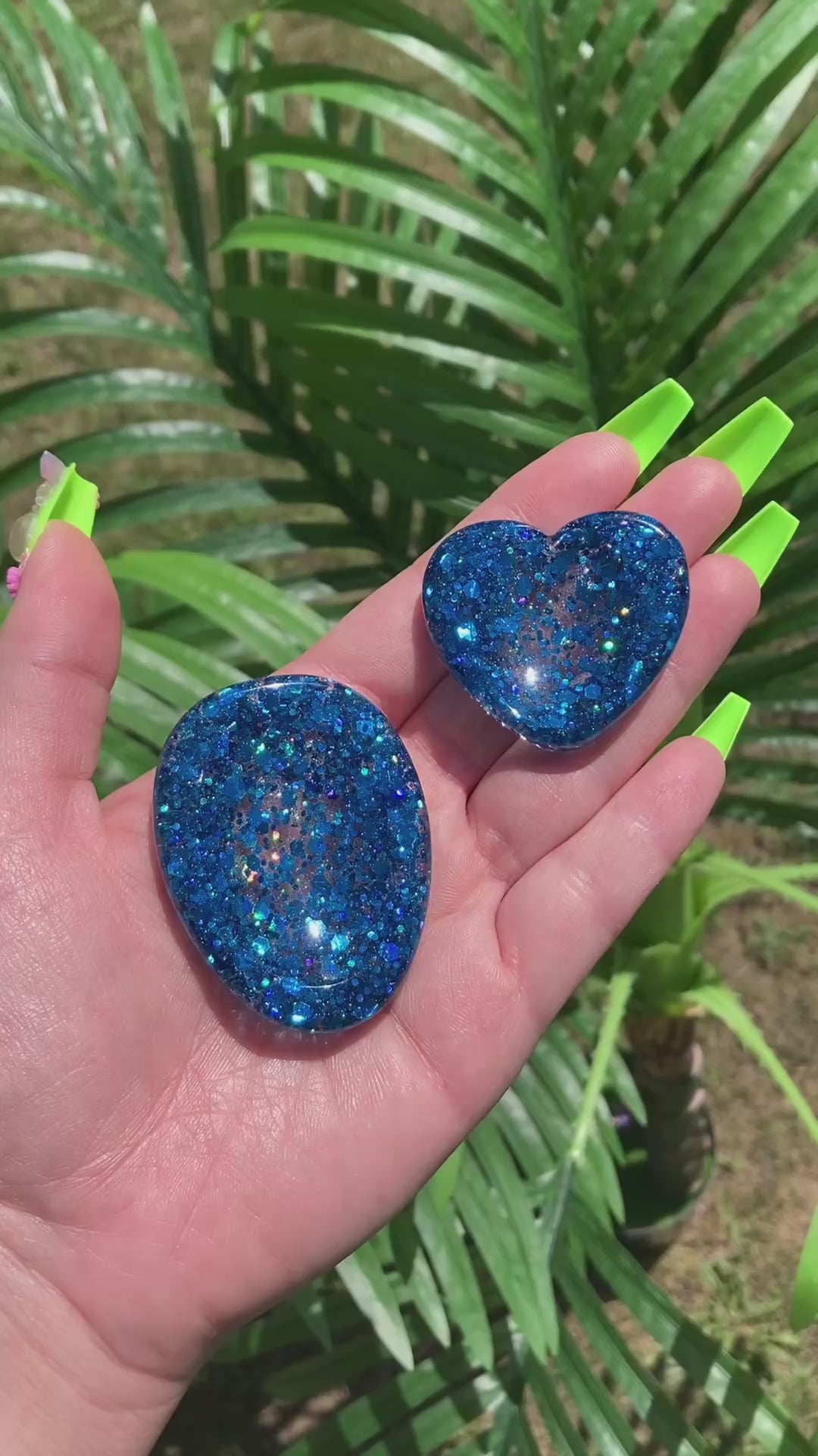 Video shows a manicured hand holding two blue sequin anxiety tokens, one heart and one oval shaped in front of an artificial plant. 